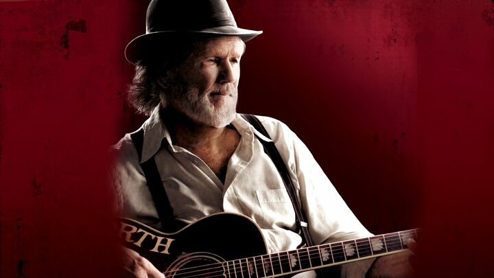 alt="Older man with guitar against red background in Bloodworth, Southern drama featuring Val Kilmer."