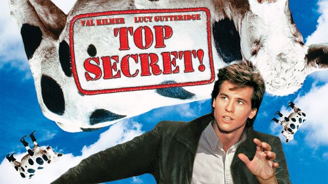 alt="Top Secret! movie poster featuring Val Kilmer in 1984 comedy classic, shown with flying cows against a blue sky"