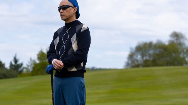alt="Ken Leung as Eric Tao stands on a golf course holding a club, wearing sunglasses and a patterned sweater."