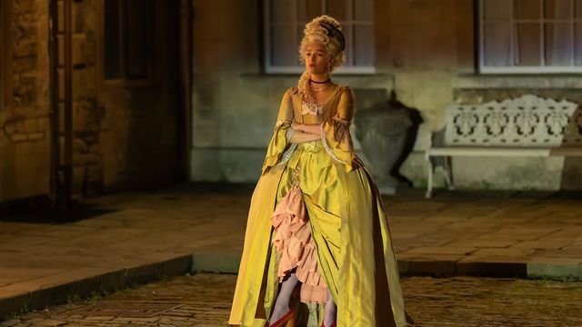 alt="Marisa Abela as Yasmin Kara-Hanani stands outdoors in an elaborate yellow period gown, arms crossed"