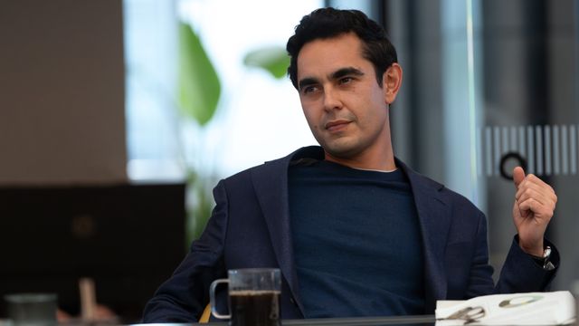 alt="Max Minghella as Whitney Halberstram sitting at a desk with coffee, gesturing while conversing to someone out of frame"